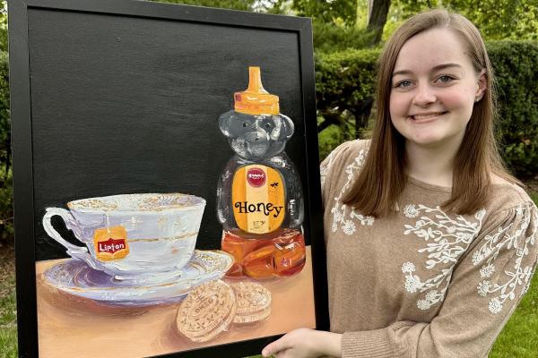 Student holds painting depicting tea, honey, and cookies;